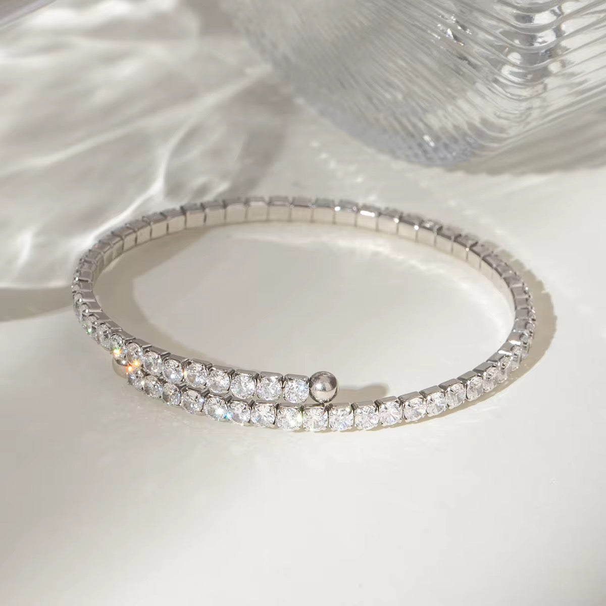 Ein flexibles Edelstahlarmband mit Goldbeschichtung und Zirkoniasteinen, präsentiert auf einer weißen Fläche mit reflektierender Lichtquelle.