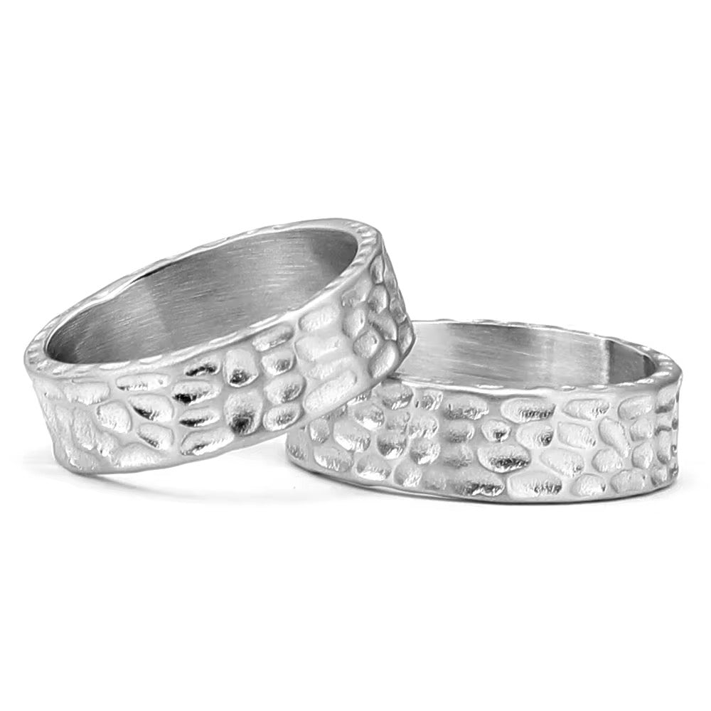 Zwei Edelstahlringe mit strukturiertem Muster, platziert auf einer dunklen, strukturierten Oberflaeche.