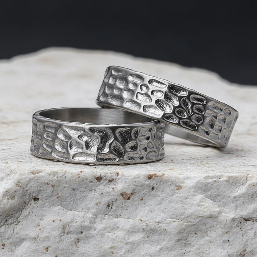 Zwei Edelstahlringe mit strukturiertem Muster, platziert auf einer dunklen, strukturierten Oberflaeche.