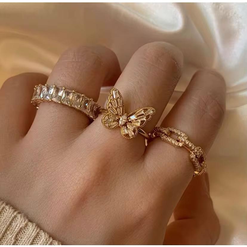 Hand mit drei goldenen Ringen mit Schmetterlingsmotiv auf einem zarten beigen Hintergrund