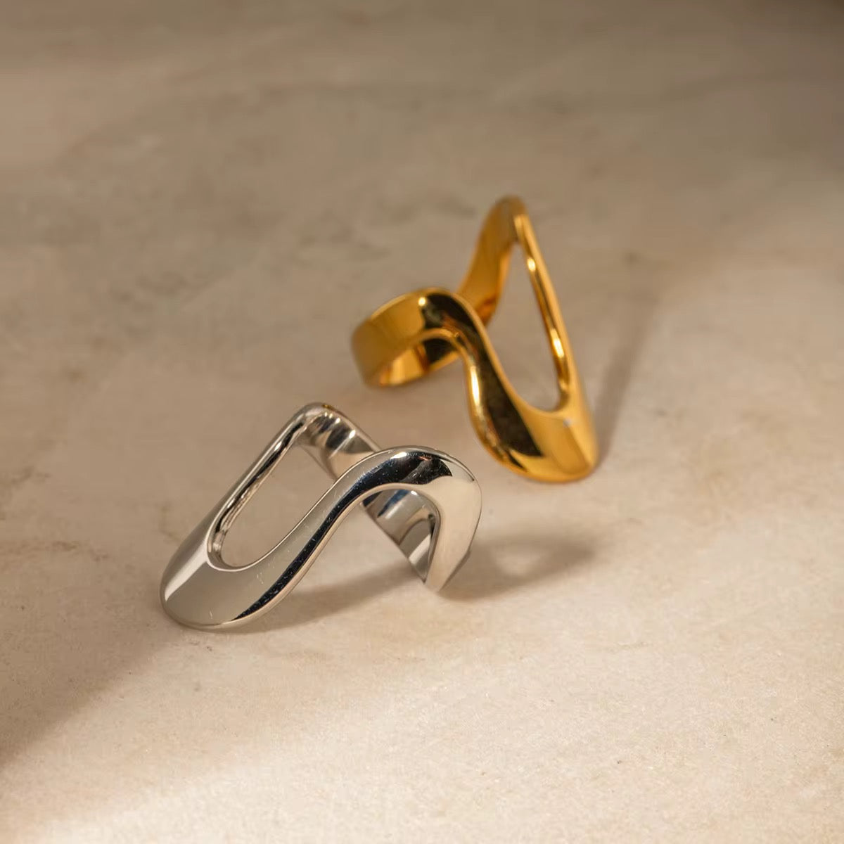 Zwei Ringe, einer mit Gold und der andere mit Silber überzogen, in unregelmäßigem Design, nebeneinander auf einer strukturierten Oberfläche platziert.