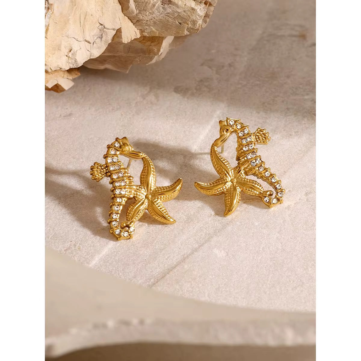 Ein Paar vergoldete Ohrringe aus Edelstahl in Form von Seepferdchen, präsentiert vor einem Naturhintergrund.
