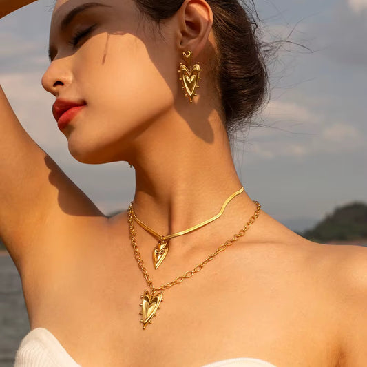 Eine Frau, die eine vergoldete Edelstahlkette mit einem herzförmigen Nietanhänger und dazu passenden Ohrringen trägt.