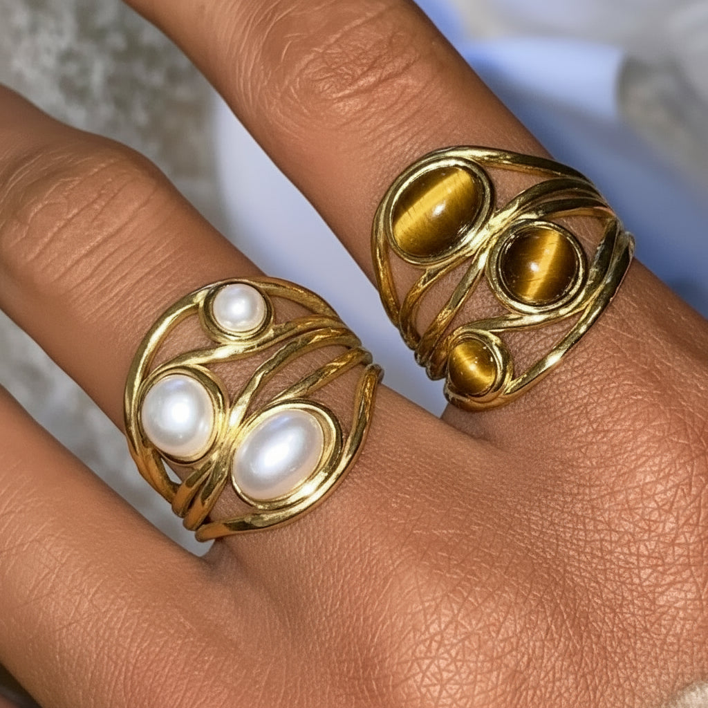 Ein vergoldeter Edelstahlring mit mehreren Bändern und Verzierungen, darunter offenbar gelbe und rosa Steine, der am Finger einer Person getragen wird.