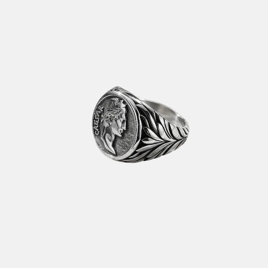 Ein versilberter Edelstahlring mit einem detaillierten Design und einer geprägten Caesar-Münze als Mittelstück, präsentiert auf weißem Untergrund.