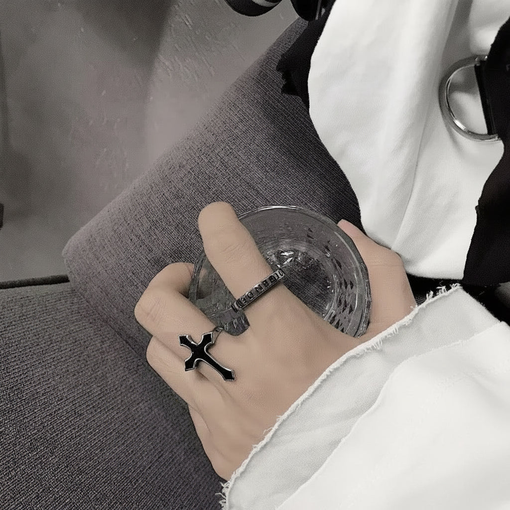 Hand mit einem schwarzen Kreuzring, die einen durchsichtigen Plastikbecher auf einer grauen Oberfläche hält.